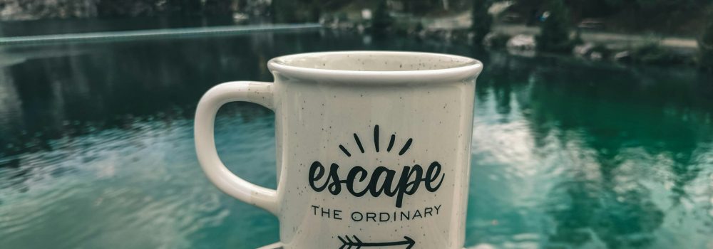 tazza con scritta "Escape the ordinary" che si affaccia su un lago di montagna. Foto di Ali Kazal su Unsplash